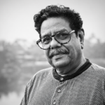 Black-and-white portrait of Gautam Siddharth, veteran writer, filmmaker, theatre practitioner, actor, and script consultant, looking toward the camera with a gentle smile. He is wearing spectacles and a textured sweater, with a blurred outdoor background. Known for his 40+ years in the Indian film industry, theatre workshops, literary contributions, and translation of over 200 films into Hindi.