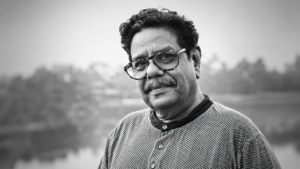 Black-and-white portrait of Gautam Siddharth, veteran writer, filmmaker, theatre practitioner, actor, and script consultant, looking toward the camera with a gentle smile. He is wearing spectacles and a textured sweater, with a blurred outdoor background. Known for his 40+ years in the Indian film industry, theatre workshops, literary contributions, and translation of over 200 films into Hindi.