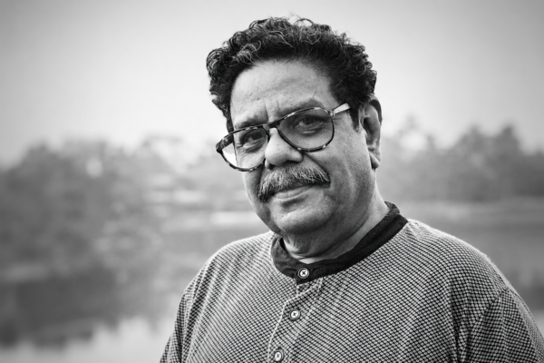 Black-and-white portrait of Gautam Siddharth, veteran writer, filmmaker, theatre practitioner, actor, and script consultant, looking toward the camera with a gentle smile. He is wearing spectacles and a textured sweater, with a blurred outdoor background. Known for his 40+ years in the Indian film industry, theatre workshops, literary contributions, and translation of over 200 films into Hindi.