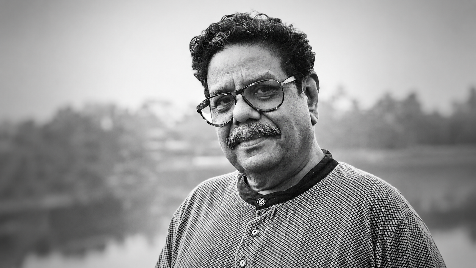 Black-and-white portrait of Gautam Siddharth, veteran writer, filmmaker, theatre practitioner, actor, and script consultant, looking toward the camera with a gentle smile. He is wearing spectacles and a textured sweater, with a blurred outdoor background. Known for his 40+ years in the Indian film industry, theatre workshops, literary contributions, and translation of over 200 films into Hindi.