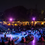 "Audience gathered at an open-air amphitheatre during a cultural and literary event at night, illuminated by vibrant stage lights with live performances, seating area, and festive stalls in the background."