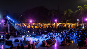"Audience gathered at an open-air amphitheatre during a cultural and literary event at night, illuminated by vibrant stage lights with live performances, seating area, and festive stalls in the background."