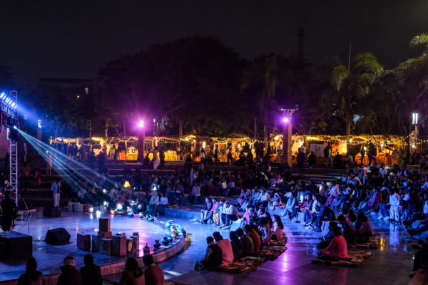 "Audience gathered at an open-air amphitheatre during a cultural and literary event at night, illuminated by vibrant stage lights with live performances, seating area, and festive stalls in the background."