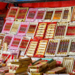 A colorful market stall displaying a wide variety of traditional Rakhi designs in red, pink, gold, and cream packaging, arranged across shelves and stacked boxes in Lucknow.