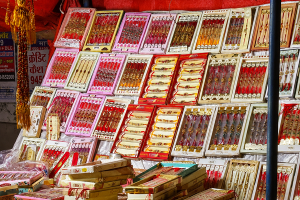 A colorful market stall displaying a wide variety of traditional Rakhi designs in red, pink, gold, and cream packaging, arranged across shelves and stacked boxes in Lucknow.