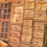 A colorful retail display featuring rows of intricately designed Rakhis arranged in decorative boxes and packaging, with traditional and floral patterns in pink, gold, red, and cream tones. The rakhi markets in Lucknow