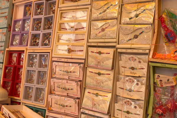 A colorful retail display featuring rows of intricately designed Rakhis arranged in decorative boxes and packaging, with traditional and floral patterns in pink, gold, red, and cream tones. The rakhi markets in Lucknow