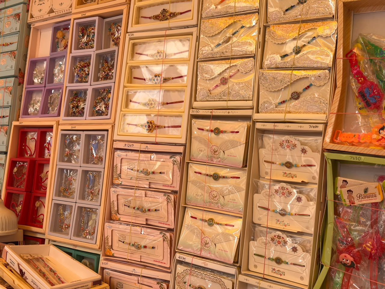 A colorful retail display featuring rows of intricately designed Rakhis arranged in decorative boxes and packaging, with traditional and floral patterns in pink, gold, red, and cream tones. The rakhi markets in Lucknow