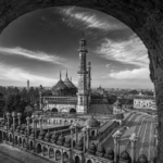 Black-and-white panoramic view of the Bara Imambara complex in Lucknow, seen through a large arched window. The image highlights the grand central mosque with its ribbed domes, soaring minarets, ornate arcades, and expansive courtyard, set against a dramatic sky with sweeping clouds. The weathered stone arch frames the historic Mughal architecture, creating depth and emphasizing the monument's scale and timeless elegance.