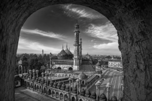 Black-and-white panoramic view of the Bara Imambara complex in Lucknow, seen through a large arched window. The image highlights the grand central mosque with its ribbed domes, soaring minarets, ornate arcades, and expansive courtyard, set against a dramatic sky with sweeping clouds. The weathered stone arch frames the historic Mughal architecture, creating depth and emphasizing the monument's scale and timeless elegance.