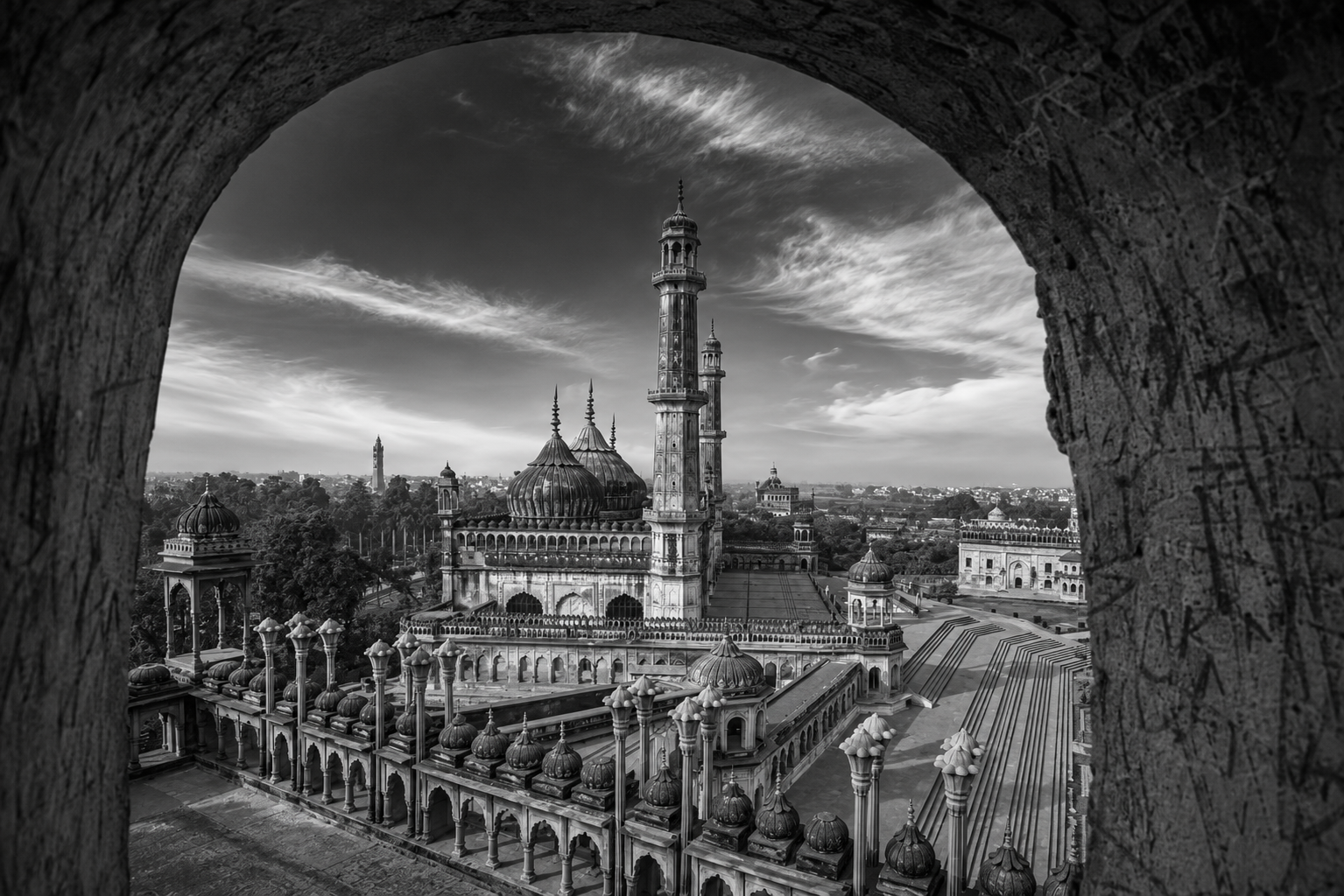 Black-and-white panoramic view of the Bara Imambara complex in Lucknow, seen through a large arched window. The image highlights the grand central mosque with its ribbed domes, soaring minarets, ornate arcades, and expansive courtyard, set against a dramatic sky with sweeping clouds. The weathered stone arch frames the historic Mughal architecture, creating depth and emphasizing the monument's scale and timeless elegance.