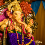 Lord Ganesha idol adorned with a golden crown, colorful garlands and traditional ornaments, seated on an ornate decorative throne with warm lighting, floral carvings, blue curtains and brass lamps in the background.