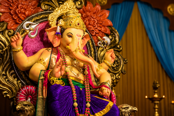 Lord Ganesha idol adorned with a golden crown, colorful garlands and traditional ornaments, seated on an ornate decorative throne with warm lighting, floral carvings, blue curtains and brass lamps in the background.