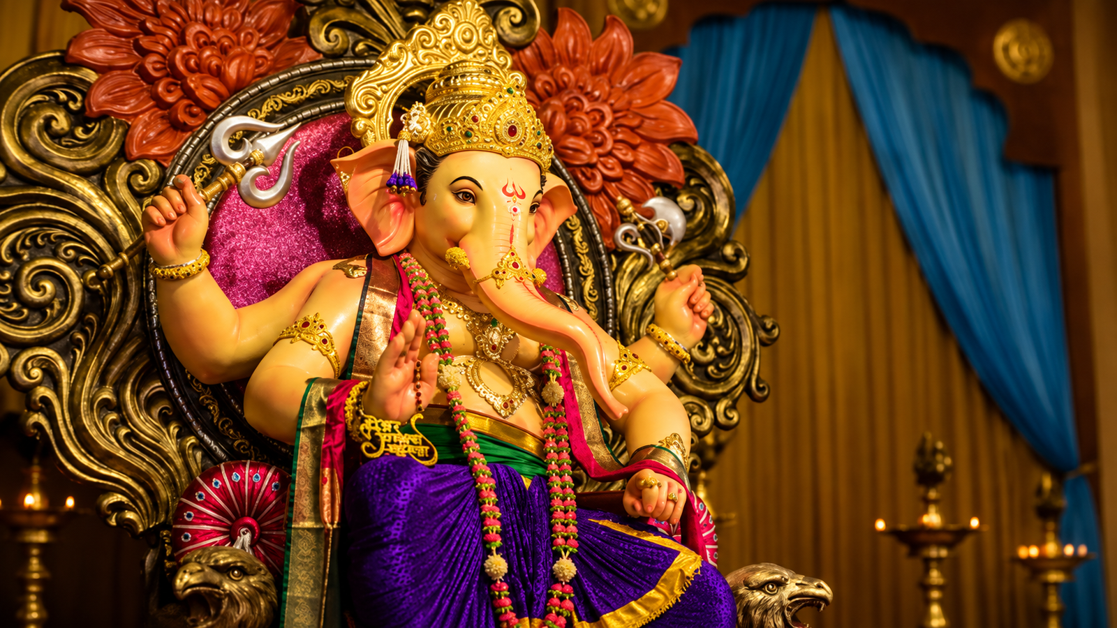 Lord Ganesha idol adorned with a golden crown, colorful garlands and traditional ornaments, seated on an ornate decorative throne with warm lighting, floral carvings, blue curtains and brass lamps in the background.