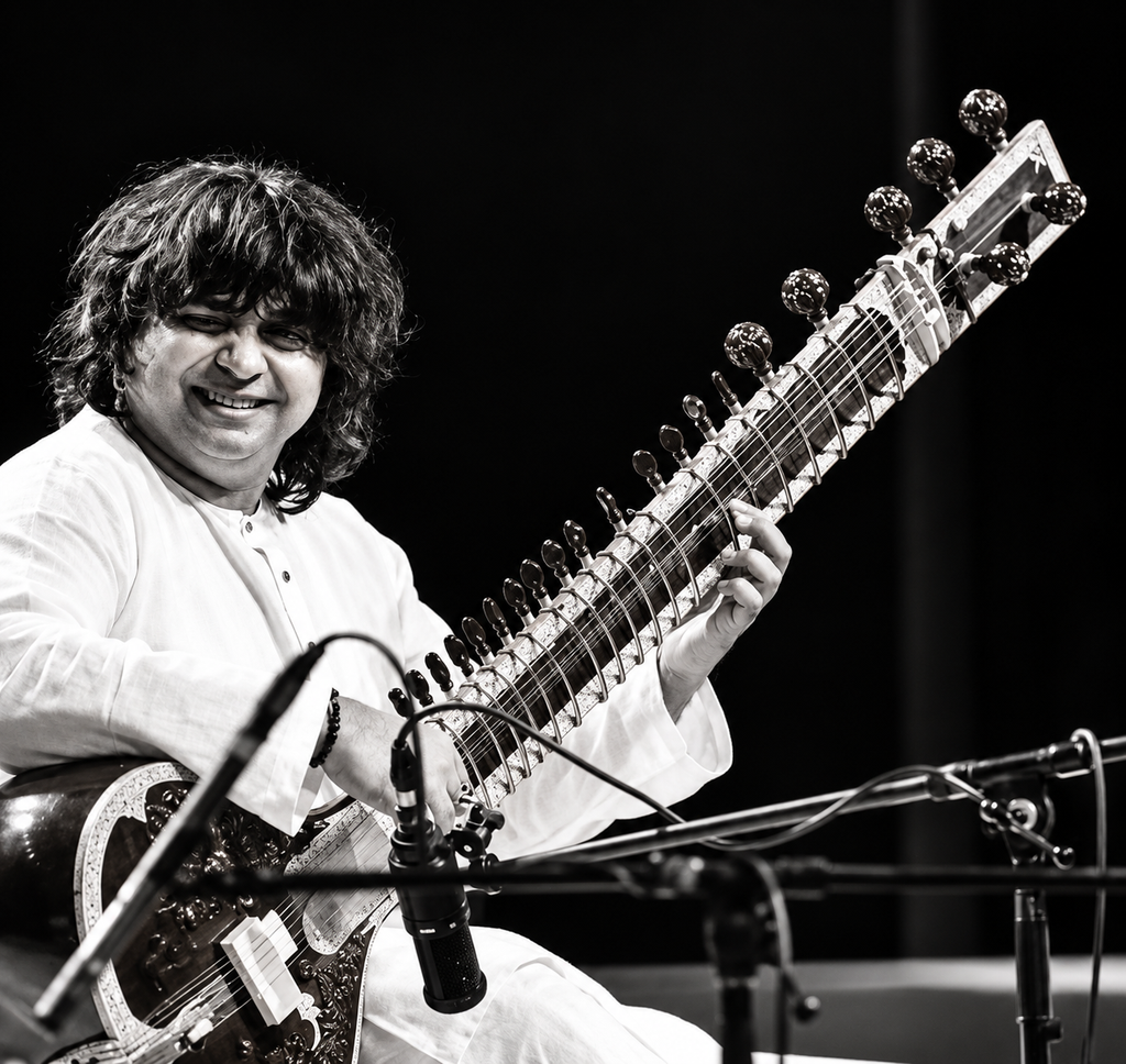 Black-and-white photograph of an Indian classical musician Niladri Kumaar performing on stage while playing an ornate sitar, with microphones and audio equipment visible in the foreground.