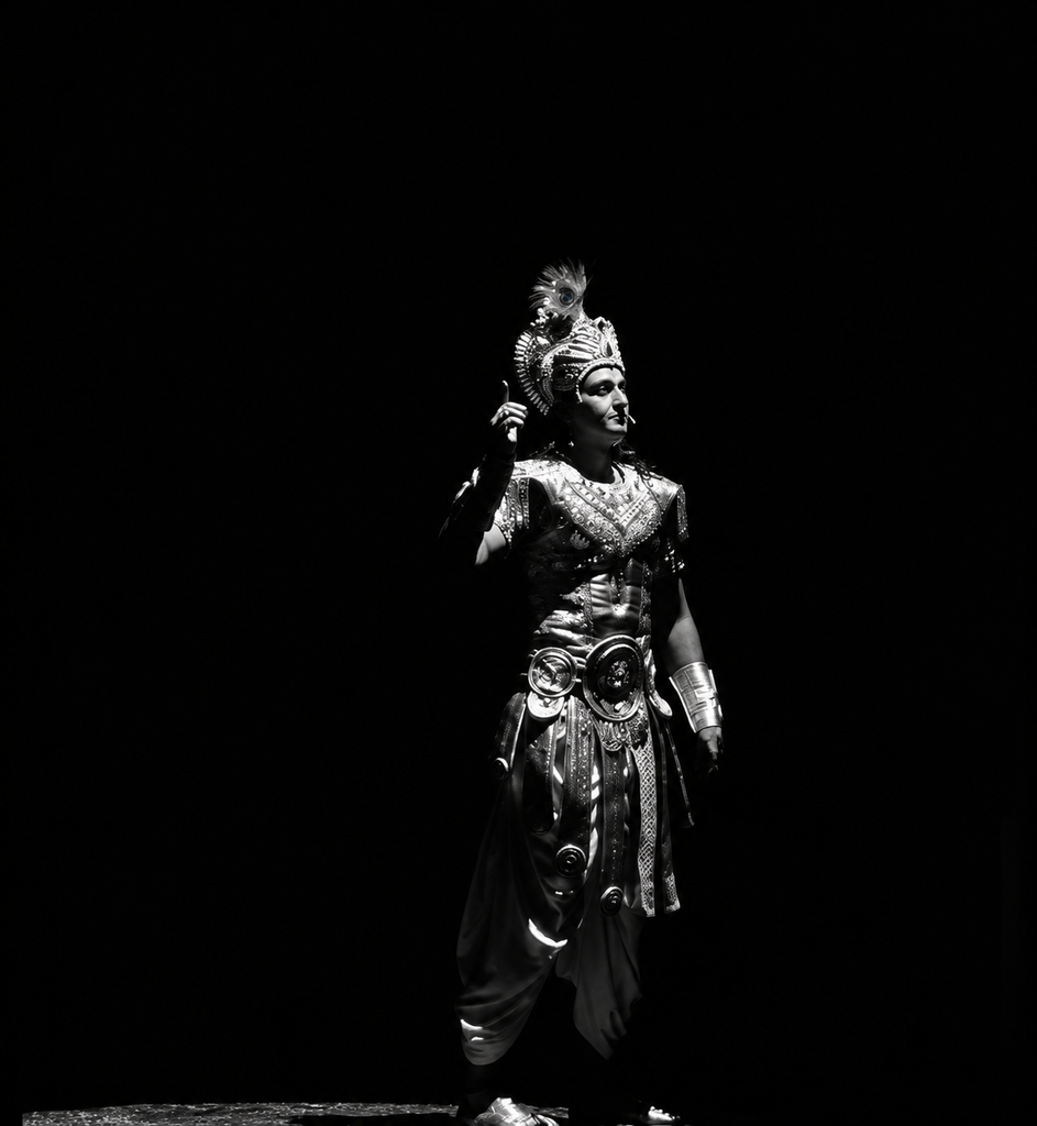 Black-and-white theatrical portrait of a male performer in an elaborate Krishna-inspired costume and ornate crown, standing under a dramatic spotlight against a dark stage background.