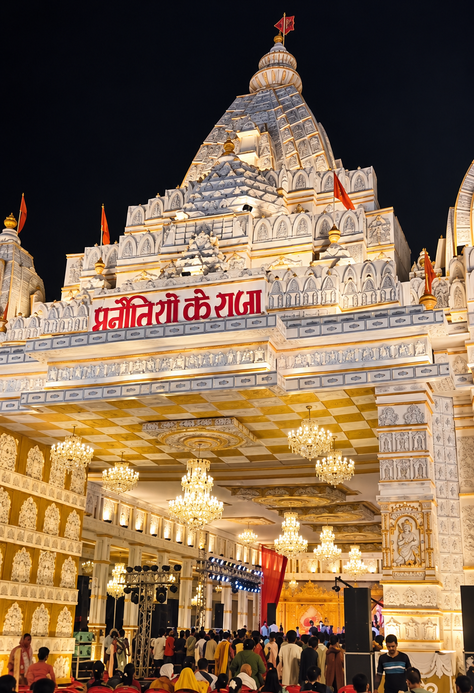 Manautiyo Ke Raja, Lucknow, beautifully illuminated at night with an ornate white-and-gold temple façade, intricate carvings, grand chandeliers, saffron flags, and devotees gathered inside.