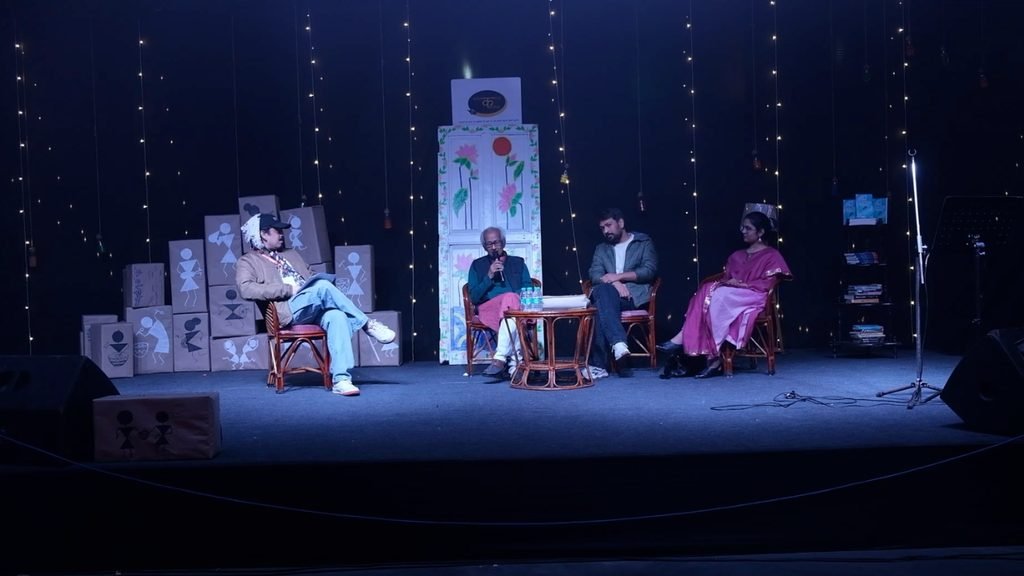 Four people seated on a decorated stage during a live literary discussion, with books, artwork, hanging lights, and a floral backdrop surrounding the panel. Naresh Saxena is the poet and writer.