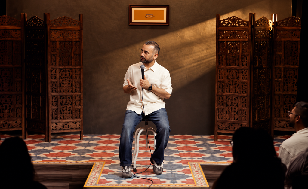 Chirag Panjwani seated on a stool on stage, holding a microphone and performing for an audience. Warm stage lighting falls across a dark backdrop with carved wooden screens, patterned flooring, and framed artwork, while audience members appear in silhouette in the foreground. This is from his specail act - Dr. Panjwani.