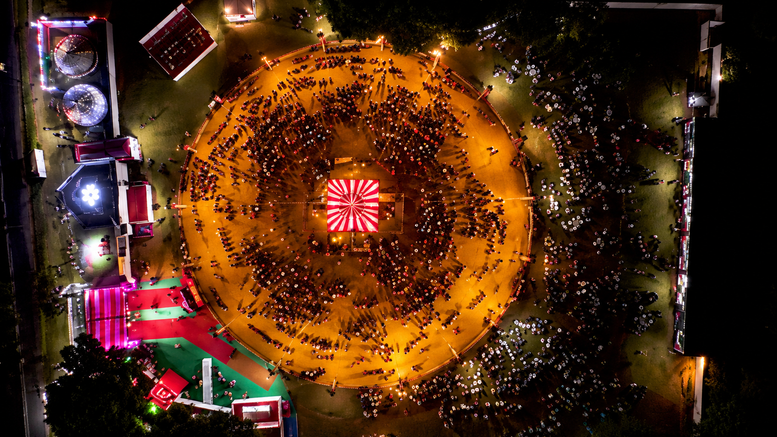 Aerial night view of Garba Nights in Lucknow, showing a large circular dance ground filled with people performing Garba around a brightly lit central stage, surrounded by colorful event areas and lights.