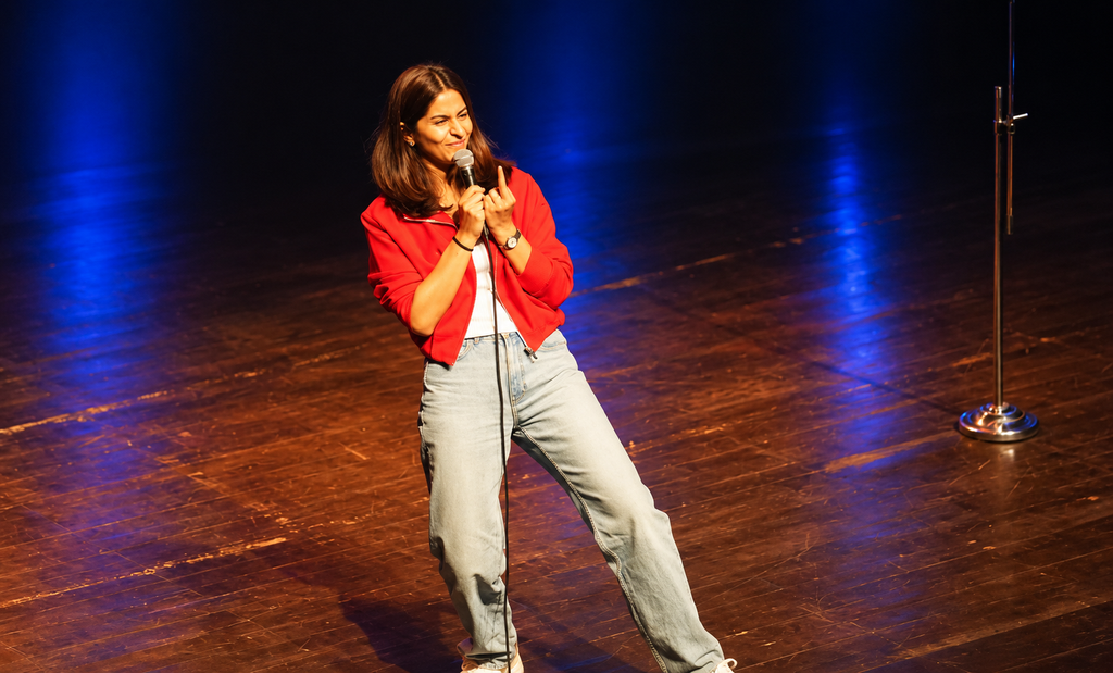 Female comedian Swati Sachdeva performing on stage with a microphone, wearing a red jacket and light jeans, under warm stage lighting with blue lights in the background.