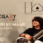 Promotional poster for Legaxy Live – Niladri Kumaar India Tour, featuring Indian classical musician Niladri Kumaar smiling while playing a sitar on the right side. The poster has a warm grey-beige textured background with subtle silhouettes of a live audience, musical elements, and architectural details. The left side prominently displays the “LEGAXY LIVE” logo, followed by the large text “NILADRI KUMAAR” and “INDIA TOUR.” The top right features the “EKKA” logo with the tagline “Enter the World of Ekka – One Sound Connects Us All,” while the bottom left includes “BLOT CANVAS.” The overall design has a vintage, artistic concert-poster aesthetic in muted grey, beige, black, cream, and brown tones.