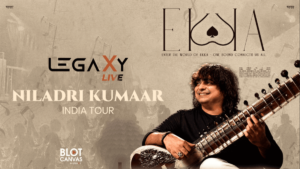 Promotional poster for Legaxy Live – Niladri Kumaar India Tour, featuring Indian classical musician Niladri Kumaar smiling while playing a sitar on the right side. The poster has a warm grey-beige textured background with subtle silhouettes of a live audience, musical elements, and architectural details. The left side prominently displays the “LEGAXY LIVE” logo, followed by the large text “NILADRI KUMAAR” and “INDIA TOUR.” The top right features the “EKKA” logo with the tagline “Enter the World of Ekka – One Sound Connects Us All,” while the bottom left includes “BLOT CANVAS.” The overall design has a vintage, artistic concert-poster aesthetic in muted grey, beige, black, cream, and brown tones.