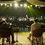 RJ Tashi and Kartik Mishra performing live at Spill The Word Fest Season 4, Day 1, on 24 October 2026, organised by Kavita Kisse Kahaniyan at Lohia Park, Lucknow, with guitars, vibrant stage lighting, and illuminated festival stalls in the background.